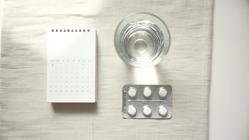 リネンの上に整然と並べられたカレンダー、丸いピルシート、水のグラスのフラットレイ。服用スケジュール管理のイメージ