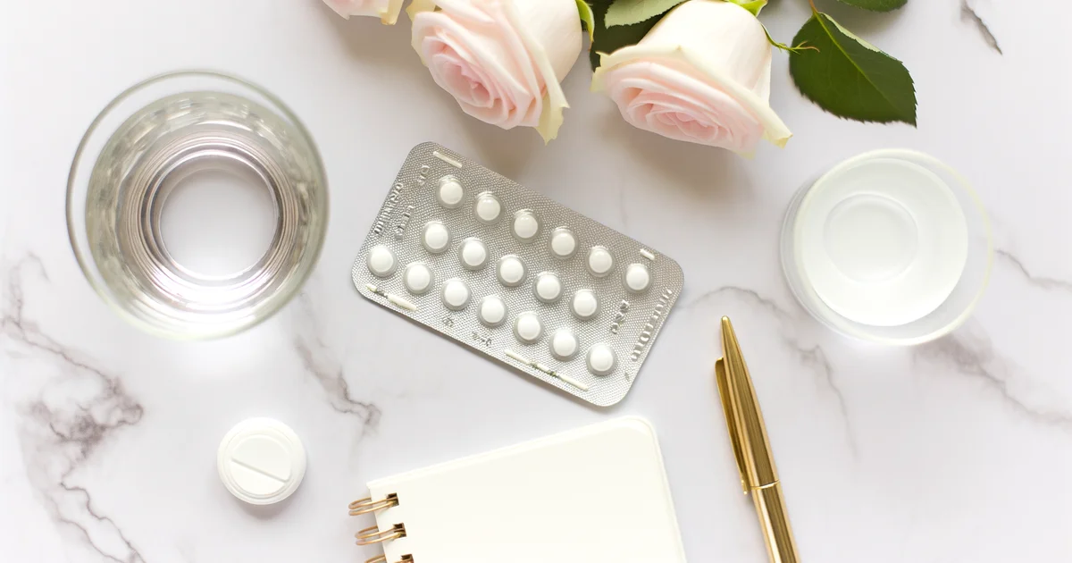 ピルのブリスターパック・水の入ったグラス・白いノート・ゴールドペン・薄いピンクのバラがマーブルデスクに並ぶフラットレイ写真